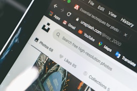 Close-up of a computer screen displaying an open web browser with multiple tabs, including a tab showing the Unsplash website. Other visible icons include web apps like Gmail, YouTube, and Maps. The search bar on the page invites users to look for free high-resolution photos.