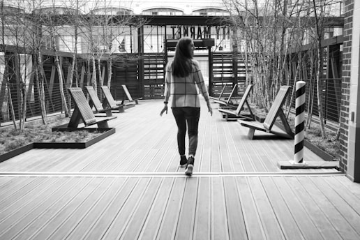 A person walks along a wooden deck with several modern, angled benches on either side. The area is surrounded by thin, leafless trees and enclosed by a contemporary architectural structure. The atmosphere is calm, with a focus on minimalist design.