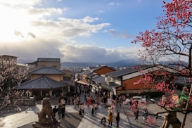 A bustling street scene in a traditional Japanese setting with a crowd of people walking along a pathway. The image features traditional buildings with tiled roofs. Cherry blossoms with bright pink flowers frame the scene on both sides, highlighting the cultural aesthetics. The sky is partly cloudy, and the sun is setting, casting warm light across the area. A statue, possibly of a mythical creature or deity, is visible in the foreground, adding a historical touch.