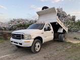 A white dump truck is parked with its bed raised, revealing a load of metal debris behind it. The truck is situated in an industrial or scrap yard area, surrounded by mounds of twisted metal and remnants of broken structures. The sky is partly cloudy, providing a backdrop to the rugged and industrious scene.