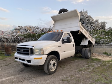 A white dump truck is parked with its bed raised, revealing a load of metal debris behind it. The truck is situated in an industrial or scrap yard area, surrounded by mounds of twisted metal and remnants of broken structures. The sky is partly cloudy, providing a backdrop to the rugged and industrious scene.