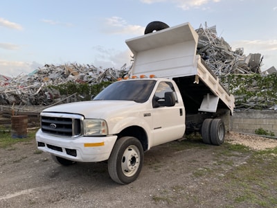 A white dump truck is parked with its bed raised, revealing a load of metal debris behind it. The truck is situated in an industrial or scrap yard area, surrounded by mounds of twisted metal and remnants of broken structures. The sky is partly cloudy, providing a backdrop to the rugged and industrious scene.