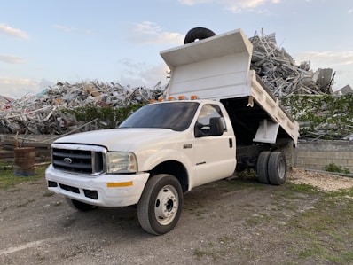 A white dump truck is parked with its bed raised, revealing a load of metal debris behind it. The truck is situated in an industrial or scrap yard area, surrounded by mounds of twisted metal and remnants of broken structures. The sky is partly cloudy, providing a backdrop to the rugged and industrious scene.