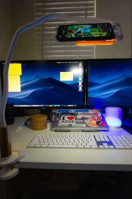A modern workspace setup featuring a dual-monitor display, keyboard, MacBook with various stickers, and a smartphone mounted on a flexible stand above the desk. A small cylindrical container, pen, lens cap, and a glowing blue light are also present on the desk. The room is dimly lit, with light coming from the screens and the blue lamp.