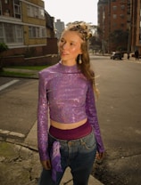 A young person with light hair styled in a creative updo, adorned with colorful hair accessories, stands on an urban street. They are wearing a sparkly, long-sleeved purple crop top, high-waisted jeans, and a purple bandana tucked into their waistband. The background includes buildings, parked cars, and other people walking in the distance.
