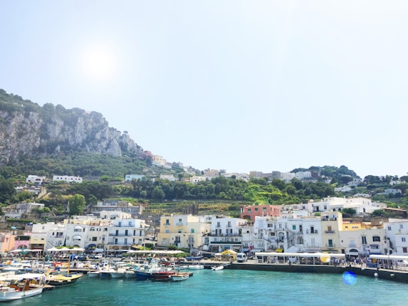 A picturesque coastal town with colorful buildings lining the waterfront, set against a backdrop of lush, green hills and rocky cliffs. Boats are docked in the vibrant blue water of the harbor, and the sun shines brightly in the clear sky.