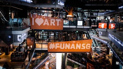 An interior scene of a modern shopping mall with multi-level walkways and escalators. Large digital billboards display messages with vibrant colors. There are glass railings, stylish flooring, and various shopfronts visible. People are casually shopping in the area.