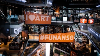 An interior scene of a modern shopping mall with multi-level walkways and escalators. Large digital billboards display messages with vibrant colors. There are glass railings, stylish flooring, and various shopfronts visible. People are casually shopping in the area.