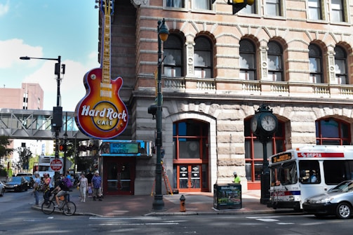 A busy urban street scene features a large guitar sign marking the entrance of a Hard Rock Cafe. The historical building facade has arched windows and detailed brickwork. Pedestrians are walking on the sidewalk, while cars and a SEPTA bus are moving along the street. A clock stands near the corner, adding a classic touch to the atmosphere.