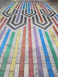 A colorful pavement design features a pattern of interlocking shapes and lines composed of rectangular tiles. Each tile row is painted in vibrant colors, including red, blue, green, yellow, and purple. The center incorporates a geometric pattern with black and white chevrons, creating a visually striking contrast.