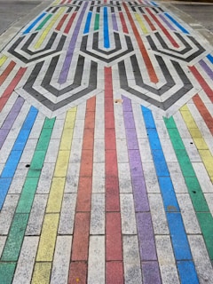 A colorful pavement design features a pattern of interlocking shapes and lines composed of rectangular tiles. Each tile row is painted in vibrant colors, including red, blue, green, yellow, and purple. The center incorporates a geometric pattern with black and white chevrons, creating a visually striking contrast.
