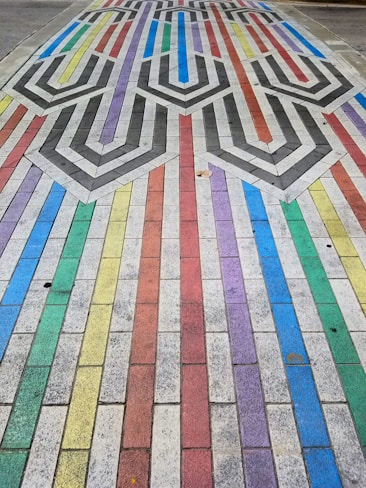 A colorful pavement design features a pattern of interlocking shapes and lines composed of rectangular tiles. Each tile row is painted in vibrant colors, including red, blue, green, yellow, and purple. The center incorporates a geometric pattern with black and white chevrons, creating a visually striking contrast.
