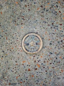 A metal survey marker is embedded in a textured concrete surface. The marker features a star in the center and has the words 'City of Austin Survey Marker' engraved on it. The concrete is speckled with various shades of gray, brown, and orange.