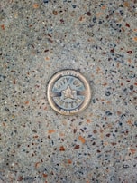 A metal survey marker is embedded in a textured concrete surface. The marker features a star in the center and has the words 'City of Austin Survey Marker' engraved on it. The concrete is speckled with various shades of gray, brown, and orange.