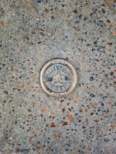 A metal survey marker is embedded in a textured concrete surface. The marker features a star in the center and has the words 'City of Austin Survey Marker' engraved on it. The concrete is speckled with various shades of gray, brown, and orange.