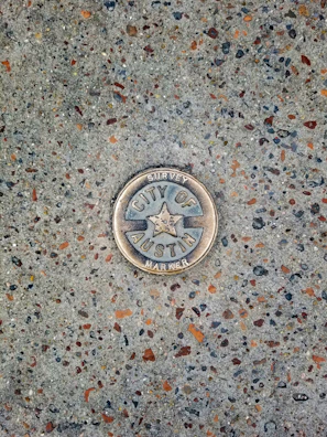 A metal survey marker is embedded in a textured concrete surface. The marker features a star in the center and has the words 'City of Austin Survey Marker' engraved on it. The concrete is speckled with various shades of gray, brown, and orange.