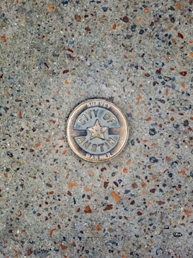 A metal survey marker is embedded in a textured concrete surface. The marker features a star in the center and has the words 'City of Austin Survey Marker' engraved on it. The concrete is speckled with various shades of gray, brown, and orange.