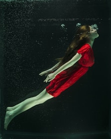 A person wearing a bright red dress is submerged underwater, appearing to be suspended in a graceful pose with bubbles rising from the face. The long hair flows freely around the head, and the limbs are extended.