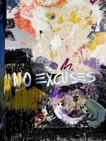 A vibrant abstract mural featuring dynamic brush strokes and splashes of paint in various colors, including yellow, purple, black, and white. The phrase 'NO EXCUSES' is prominently painted in bold white letters across the artwork, contrasted against darker hues. The composition is chaotic and expressive, with a mixture of forms and textures.