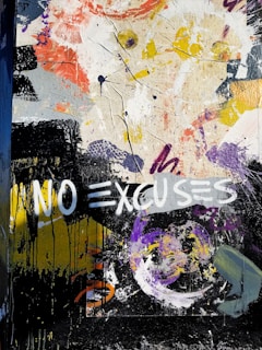 A vibrant abstract mural featuring dynamic brush strokes and splashes of paint in various colors, including yellow, purple, black, and white. The phrase 'NO EXCUSES' is prominently painted in bold white letters across the artwork, contrasted against darker hues. The composition is chaotic and expressive, with a mixture of forms and textures.