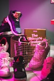 A collection of sneakers arranged around a wire basket, with a book titled 'The Ultimate Sneaker Book' inside it. The sneakers vary in colors and designs, positioned on a tiled floor. A pink and purple lighting effect creates a vibrant atmosphere.