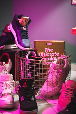 A collection of sneakers arranged around a wire basket, with a book titled 'The Ultimate Sneaker Book' inside it. The sneakers vary in colors and designs, positioned on a tiled floor. A pink and purple lighting effect creates a vibrant atmosphere.