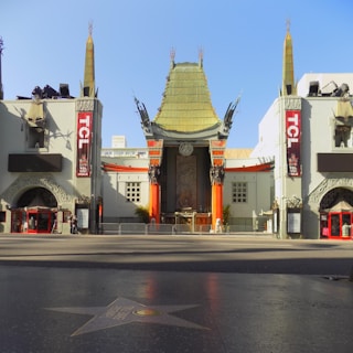 An iconic theater with an elaborate, pagoda-style roof structure, featuring intricate architectural details. The theater is flanked by two vertical signs with red lettering. The front courtyard includes the famous Hollywood Walk of Fame, visible in the foreground with a star embedded in the pavement.