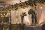 An elegantly decorated banquet setting featuring tall arrangements of white and cream flowers on stands, with cascading flowers overhead. Warm lighting from hanging bulbs adds a soft glow to the scene, complementing the flowing draped fabrics in the background and on the tables.