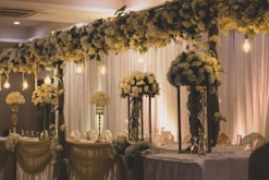An elegantly decorated banquet setting featuring tall arrangements of white and cream flowers on stands, with cascading flowers overhead. Warm lighting from hanging bulbs adds a soft glow to the scene, complementing the flowing draped fabrics in the background and on the tables.