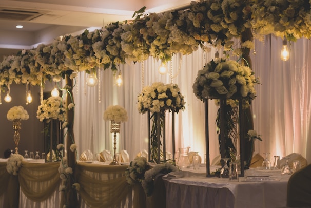 An elegantly decorated banquet setting featuring tall arrangements of white and cream flowers on stands, with cascading flowers overhead. Warm lighting from hanging bulbs adds a soft glow to the scene, complementing the flowing draped fabrics in the background and on the tables.