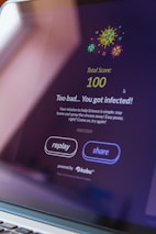 A computer screen displays a game interface with colorful virus illustrations at the top. The text reads 'Total Score: 100' followed by 'Too bad... You got infected!' The interface includes buttons labeled 'replay' and 'share' and mentions it's powered by 'kobu'.