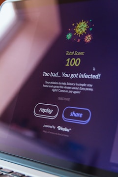 A computer screen displays a game interface with colorful virus illustrations at the top. The text reads 'Total Score: 100' followed by 'Too bad... You got infected!' The interface includes buttons labeled 'replay' and 'share' and mentions it's powered by 'kobu'.