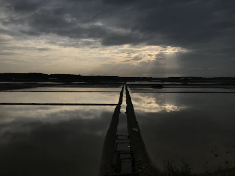 An expansive landscape features a series of flat, reflective water surfaces likely used for agriculture or salt production. The sky is filled with thick, dark clouds with a subtle break allowing sun rays to pierce through, creating a dramatic contrast in the lighting. A narrow, elevated path divides the water surfaces.