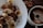 Slices of banana are generously sprinkled with cocoa powder on a white plate, accompanied by a cup containing a dark brown beverage. Cocoa beans are scattered on a wooden surface next to the plate, suggesting themes of chocolate and coffee.