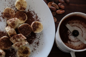Slices of banana are generously sprinkled with cocoa powder on a white plate, accompanied by a cup containing a dark brown beverage. Cocoa beans are scattered on a wooden surface next to the plate, suggesting themes of chocolate and coffee.