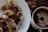 Slices of banana are generously sprinkled with cocoa powder on a white plate, accompanied by a cup containing a dark brown beverage. Cocoa beans are scattered on a wooden surface next to the plate, suggesting themes of chocolate and coffee.