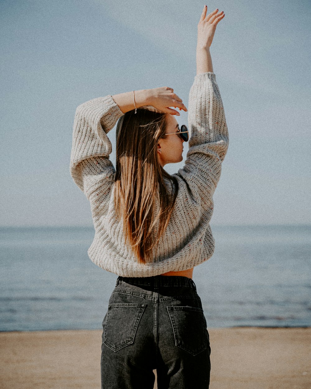 woman in white sweater and blue denim jeans standing on beach shore during daytime