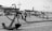 A black and white image of a maritime scene featuring a large anchor in the foreground. The scene includes a docked ship with visible smokestacks and nautical fixtures like chains and bollards. The scene is set on a cobblestone surface with lampposts and a maritime museum or harbor area in the background.