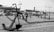 A black and white image of a maritime scene featuring a large anchor in the foreground. The scene includes a docked ship with visible smokestacks and nautical fixtures like chains and bollards. The scene is set on a cobblestone surface with lampposts and a maritime museum or harbor area in the background.
