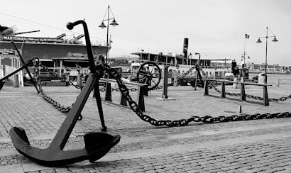 A black and white image of a maritime scene featuring a large anchor in the foreground. The scene includes a docked ship with visible smokestacks and nautical fixtures like chains and bollards. The scene is set on a cobblestone surface with lampposts and a maritime museum or harbor area in the background.