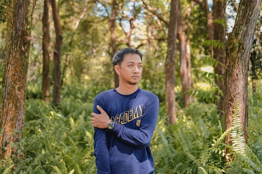 A person wearing a navy blue shirt with the word 'ACADEMY' is standing in a lush, green forest. The individual is holding their left arm with their right hand, and their expression appears contemplative. Surrounding them are tall trees and abundant ferns, creating a serene natural environment.
