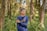 A person wearing a navy blue shirt with the word 'ACADEMY' is standing in a lush, green forest. The individual is holding their left arm with their right hand, and their expression appears contemplative. Surrounding them are tall trees and abundant ferns, creating a serene natural environment.