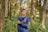 A person wearing a navy blue shirt with the word 'ACADEMY' is standing in a lush, green forest. The individual is holding their left arm with their right hand, and their expression appears contemplative. Surrounding them are tall trees and abundant ferns, creating a serene natural environment.