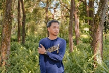 A person wearing a navy blue shirt with the word 'ACADEMY' is standing in a lush, green forest. The individual is holding their left arm with their right hand, and their expression appears contemplative. Surrounding them are tall trees and abundant ferns, creating a serene natural environment.