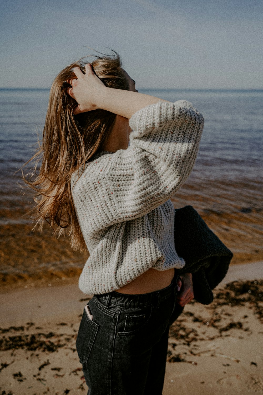 woman in white knit sweater and blue denim jeans standing on beach shore during daytime