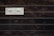 A wooden wall with horizontal planks features a vintage-style rectangular sign displaying the word 'Welcome' in bold black letters. The background of the sign is off-white with a decorative border in a rusty metal frame.