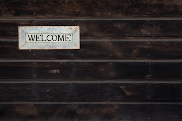 A wooden wall with horizontal planks features a vintage-style rectangular sign displaying the word 'Welcome' in bold black letters. The background of the sign is off-white with a decorative border in a rusty metal frame.