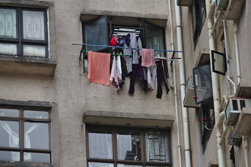Clothes are hanging to dry on a metal rod installed outside a window of an apartment building. The facade of the building is constructed with a beige textured material, and nearby windows are partially open. Multiple garments, including a striped shirt and various colorful clothes, are visible on the rod.
