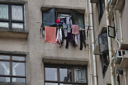 Clothes are hanging to dry on a metal rod installed outside a window of an apartment building. The facade of the building is constructed with a beige textured material, and nearby windows are partially open. Multiple garments, including a striped shirt and various colorful clothes, are visible on the rod.