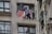 Clothes are hanging to dry on a metal rod installed outside a window of an apartment building. The facade of the building is constructed with a beige textured material, and nearby windows are partially open. Multiple garments, including a striped shirt and various colorful clothes, are visible on the rod.